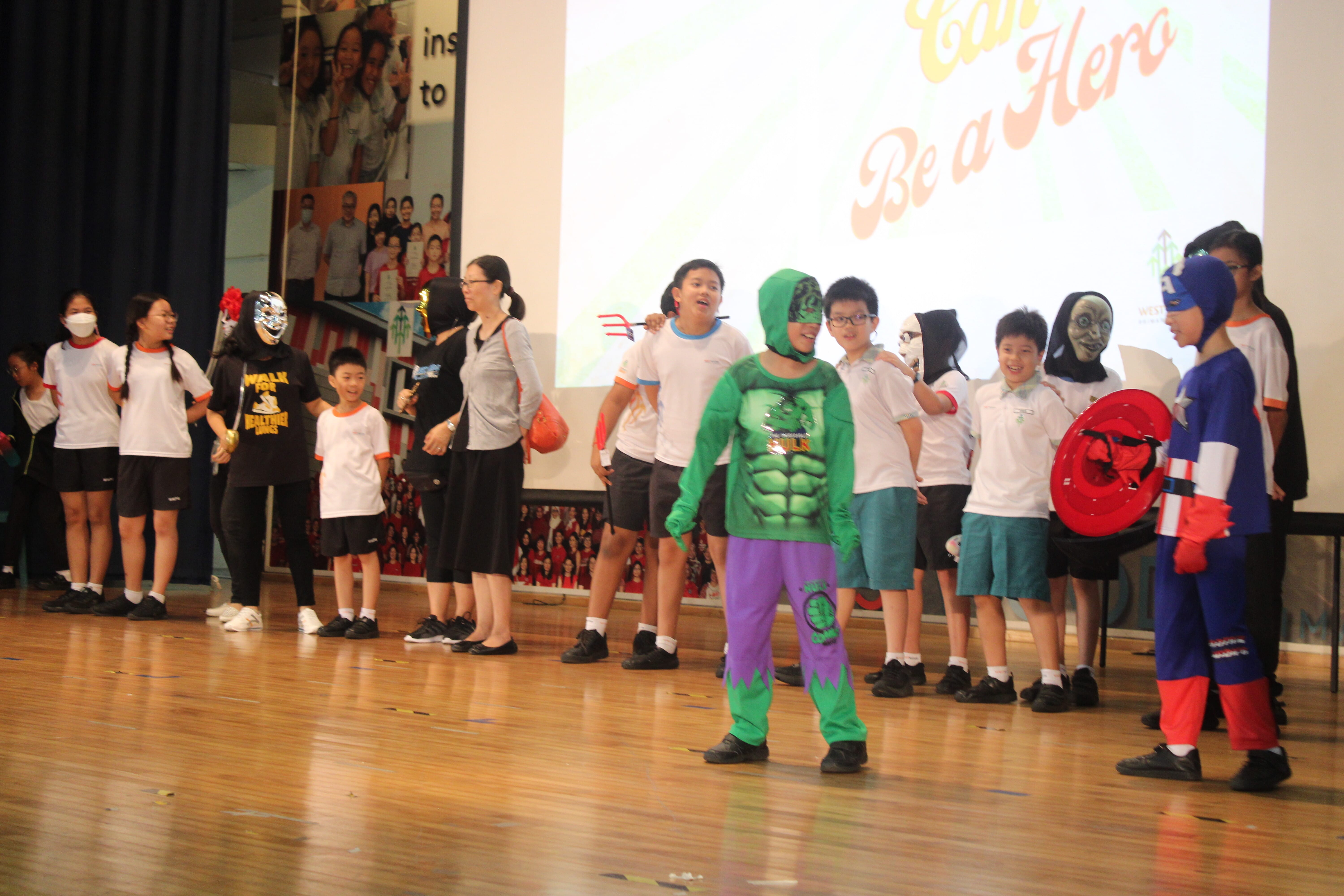 Children on a stage, some in superhero costumes like Hulk and Captain America, with a "Be a Hero" projected screen behind them.